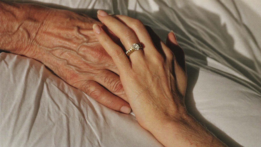 An older hand and a younger hand resting together on white linen, a diamond ring catching the morning light
