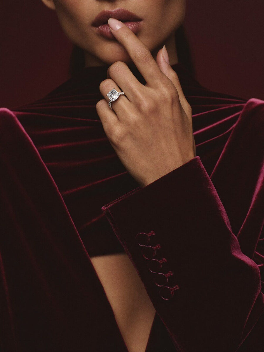 A hand in oxblood velvet wearing a diamond ring, fingertip at her lips