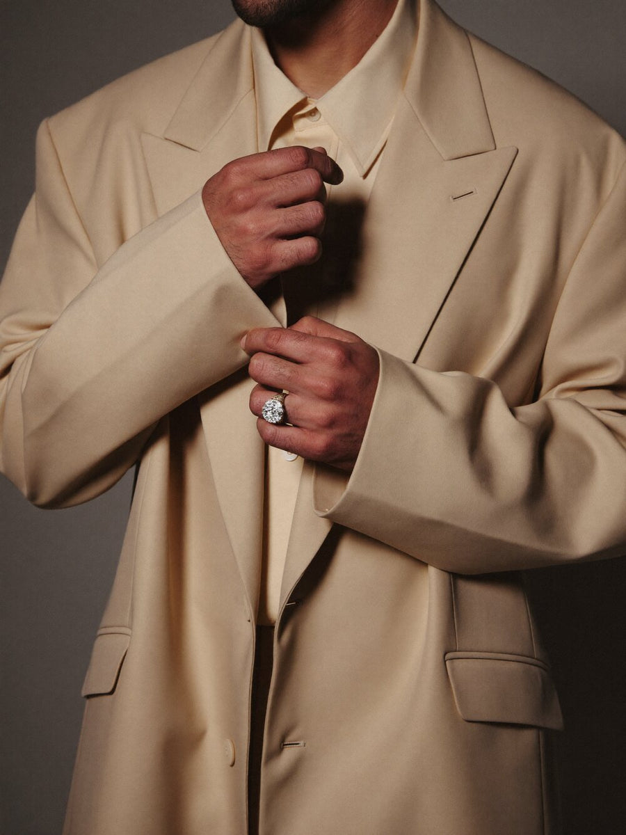 A man in cream tailoring adjusting his cuff, a diamond ring on his finger